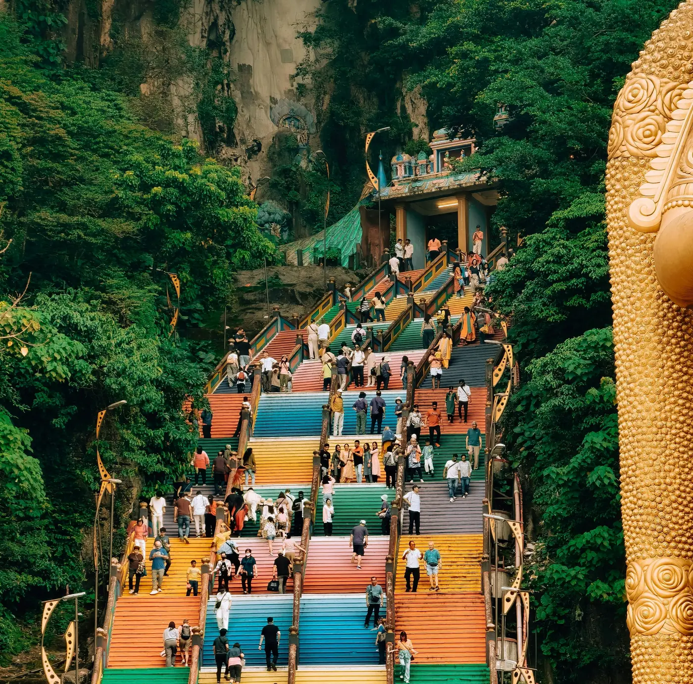 Las escaleras arcoíris de Batu Caves llenas de gente subiendo, con la estatua dorada de Murugan al costado